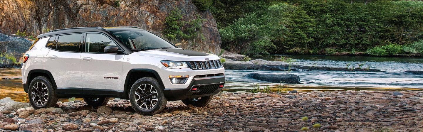A white 2021 Jeep Compass Trailhawk parked on a rocky trail, next to a stream in the woods.