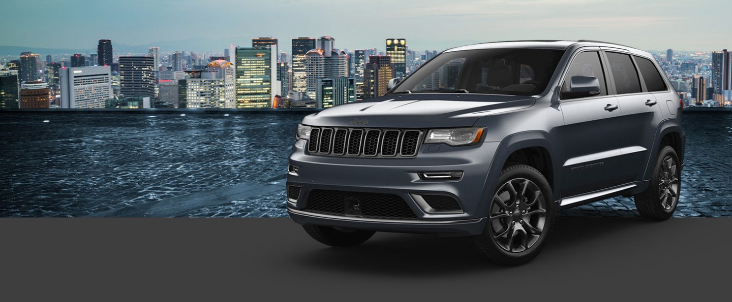 A gray 2021 Jeep Grand Cherokee High Altitude in the foreground, with a cityscape at sunrise in the background.