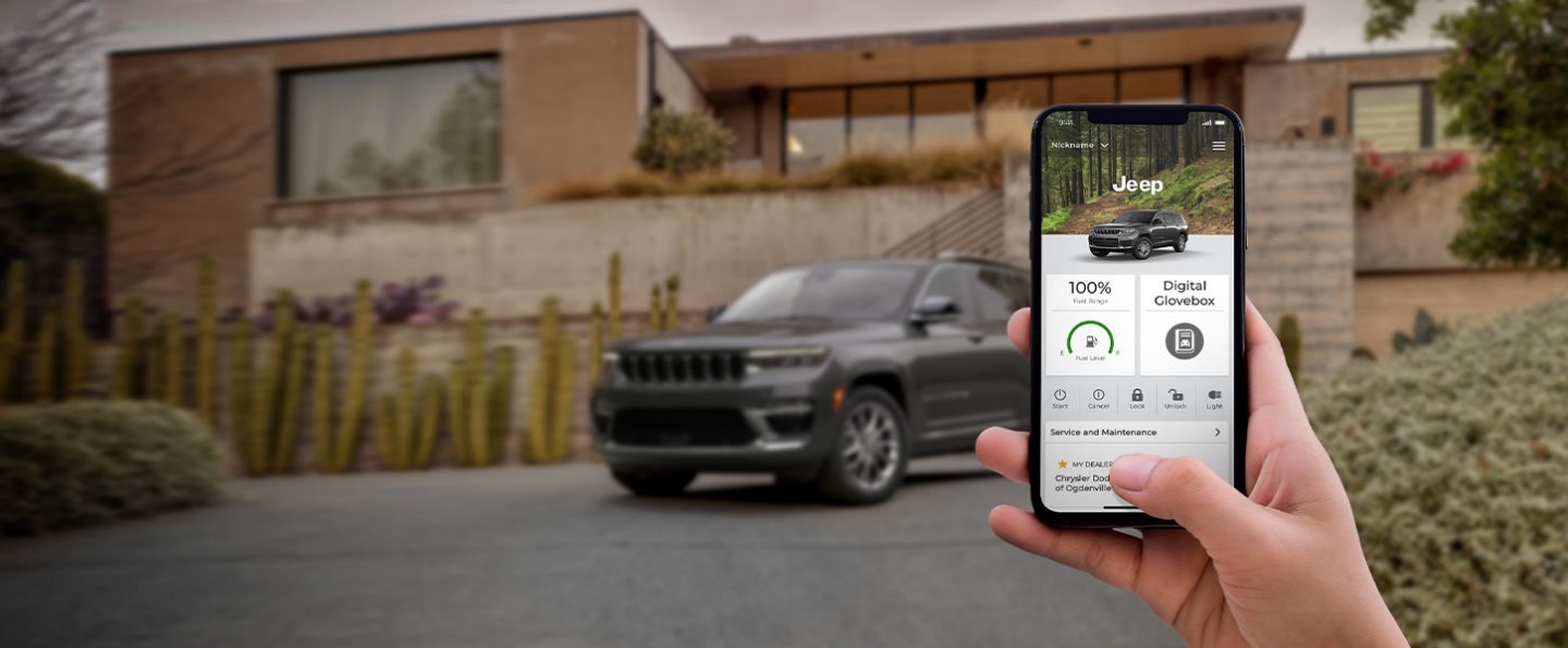 In the foreground, a hand holding a smartphone displaying the Jeep Connect app. In the background, a 2022 Jeep Grand Cherokee L.