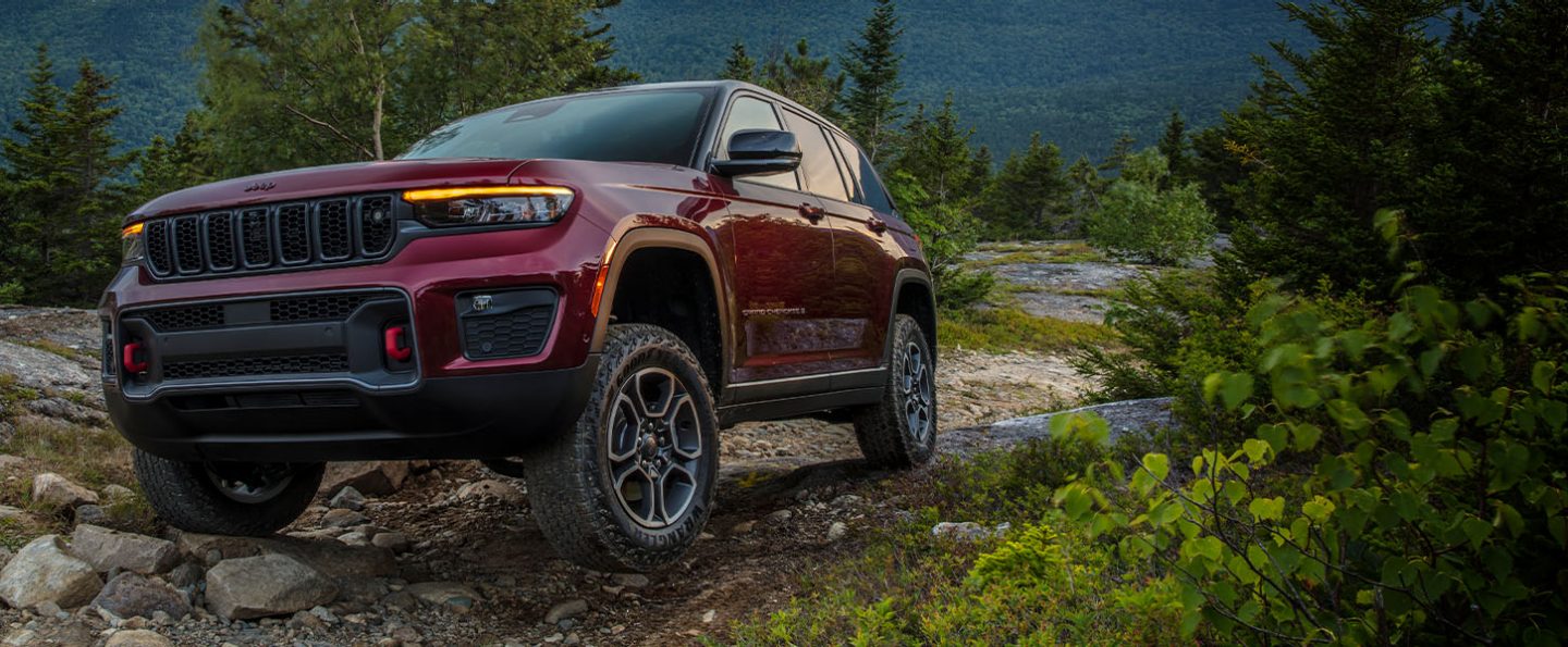 A 2022 Jeep Grand Cherokee Trailhawk parked on a rocky incline at an angle that highlights the vehicle's available ground clearance. Forested hills are visible in the background.