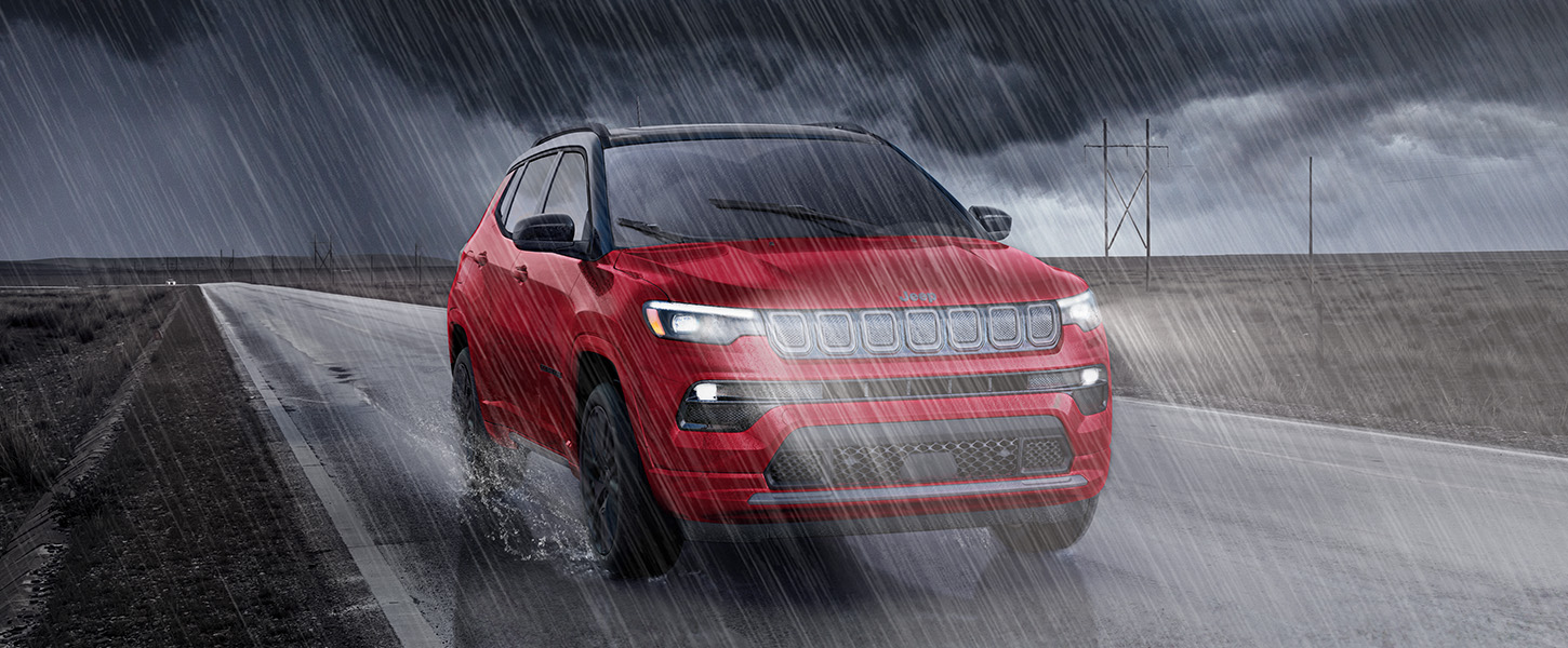 A slider that when pulled to the right reveals a 2023 Jeep Compass Limited being driven on a highway between two open fields at sunset; and when pulled to the left reveals the same 2023 Jeep Compass Limited being driven through a dark, heavy rainstorm on a highway between two open fields.