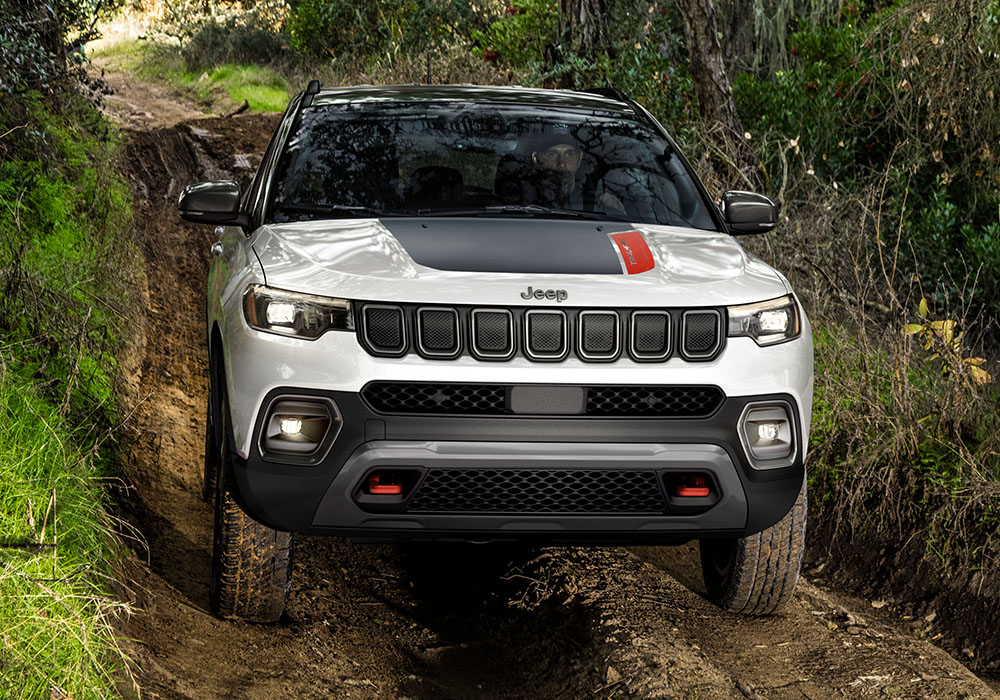 A head-on view of a 2023 Jeep Compass Trailhawk being driven off-road through a narrow trail with thick growth on both sides.