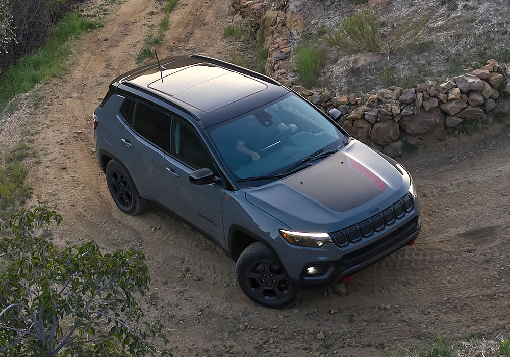 A 2023 Jeep Compass Trailhawk being driven on a dirt road hill beside a stone fence.
