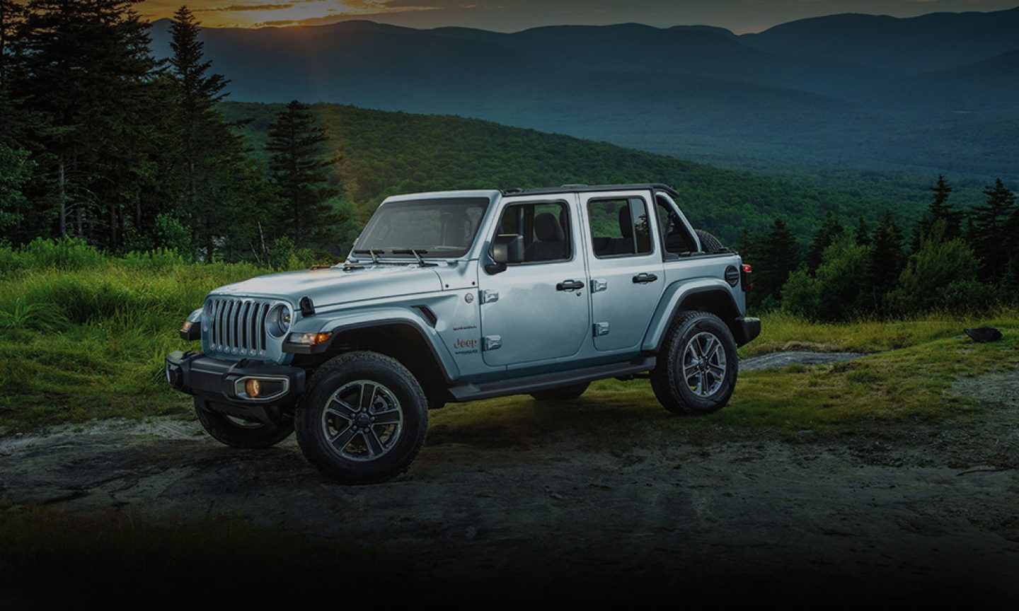 The 2023 Jeep Wrangler Sahara parked off-road on a patch of bare earth surrounded by grass and trees.