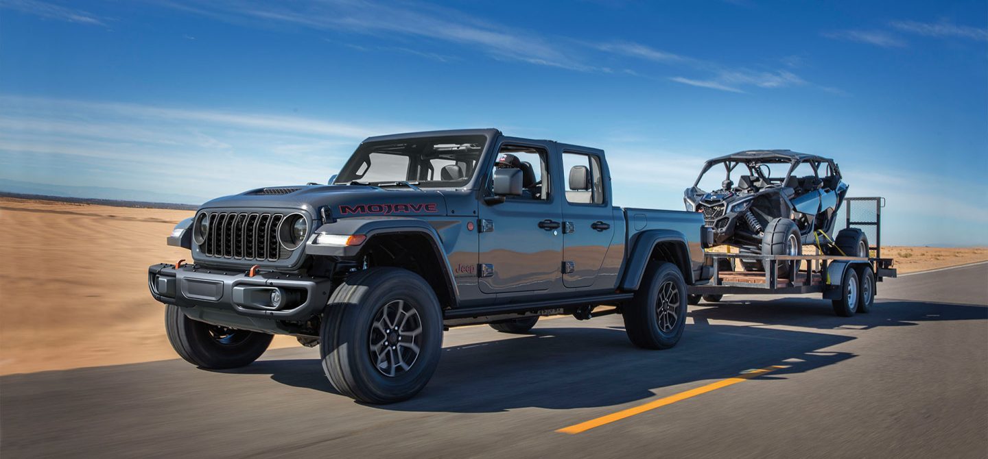 A gray 2025 Jeep Gladiator Mojave X with its roof removed, being driven on a highway in the desert, towing a dune buggy.