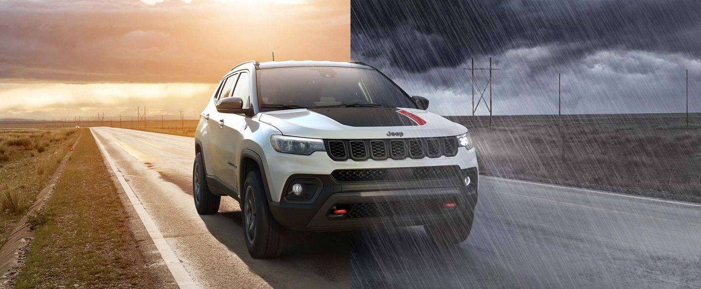 A split-screen of a white 2026 Jeep Compass Trailhawk with black hood insert, traveling on a highway between two open fields. On the left, it is a clear day at sunset; and on the right, a dark, cloudy sky pours heavy rain on the highway.