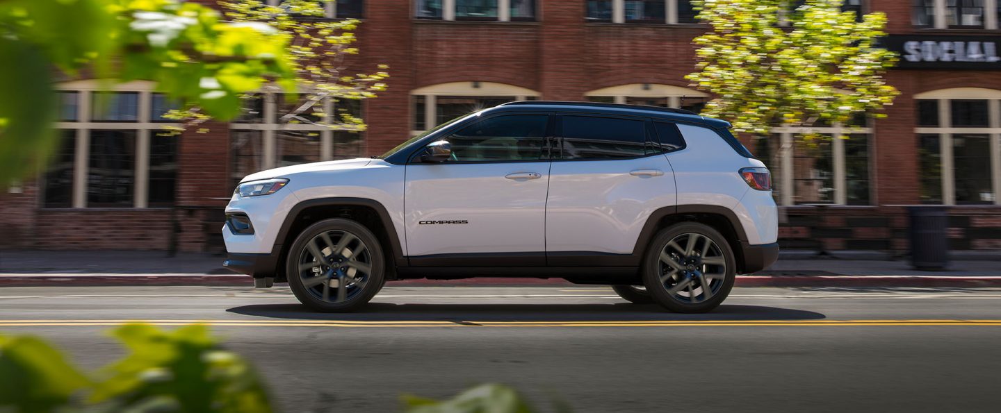 A driver-side profile of a white 2026 Jeep Compass Limited Altitude with a black hood insert, traveling on a city street. The background is blurred to indicate the vehicle is in motion.
