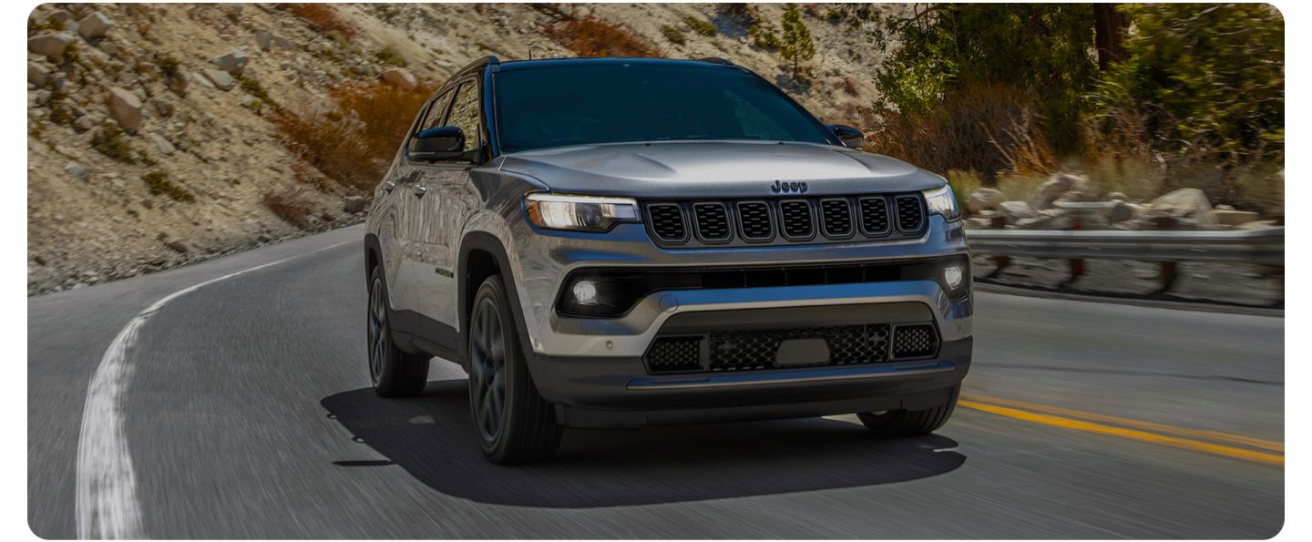 A front angle of a silver 2026 Jeep Compass Limited Altitude traveling on a winding mountain highway.