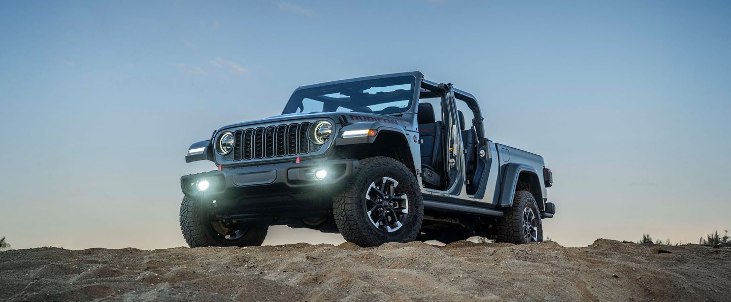 A gray 2026 Jeep Gladiator Rubicon X with its doors and top removed and its headlamps and fog lamps on, parked on the top of a rocky hill.