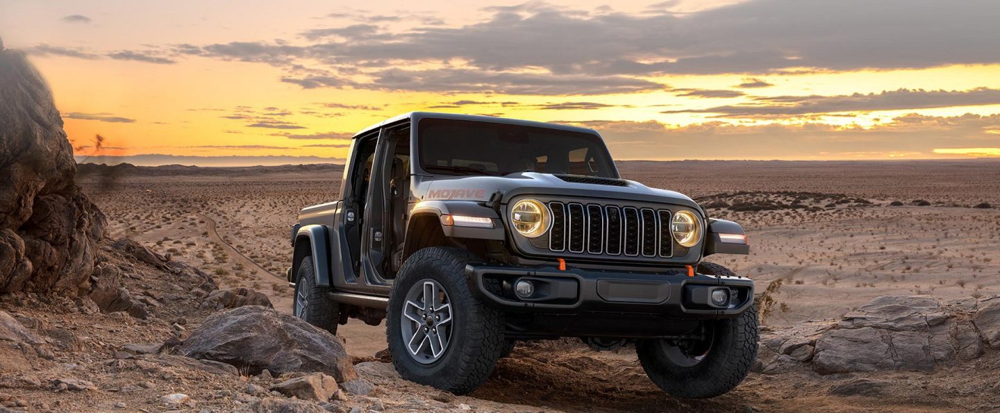 A gray 2026 Jeep Gladiator Mojave with its doors removed, ascending a hill in the desert at sunset.