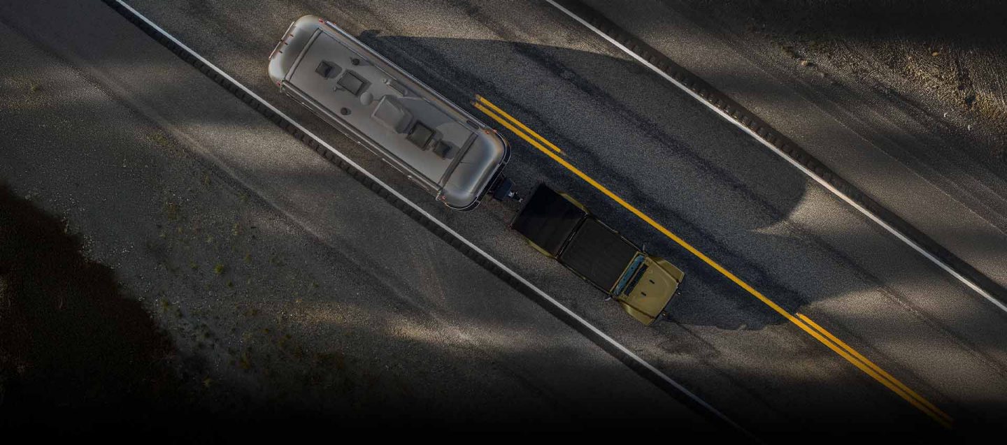 An overhead angle of a olive green 2026 Jeep Gladiator traveling down a highway, towing a large travel trailer.