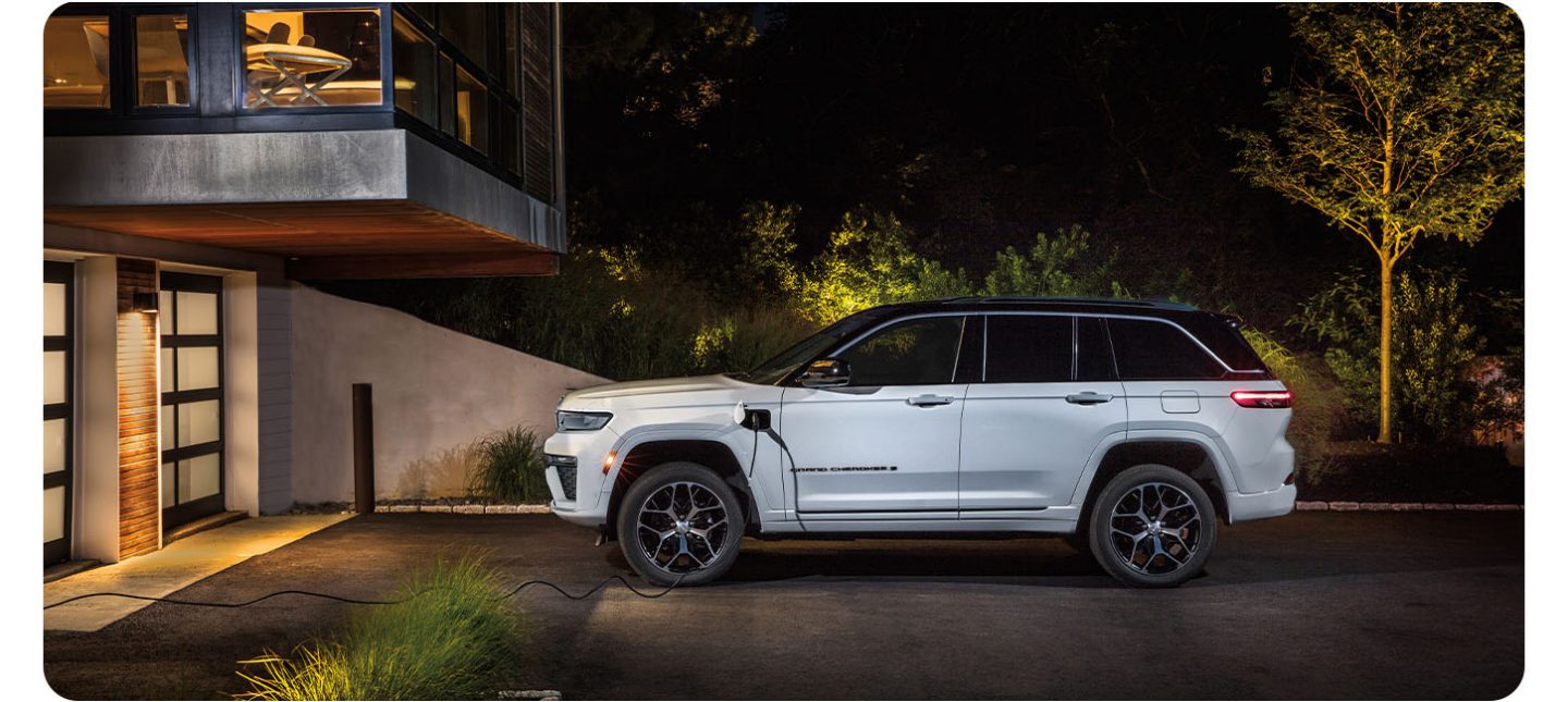 A driver-side profile of a white 2026 Jeep Grand Cherokee Summit 4xe parked in the driveway of a large home, with a charging cord plugged into the vehicle's charging port.