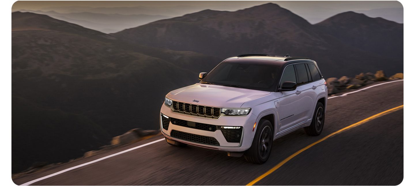A white 2026 Jeep Grand Cherokee Summit 4xe traveling down a mountain road at dusk.