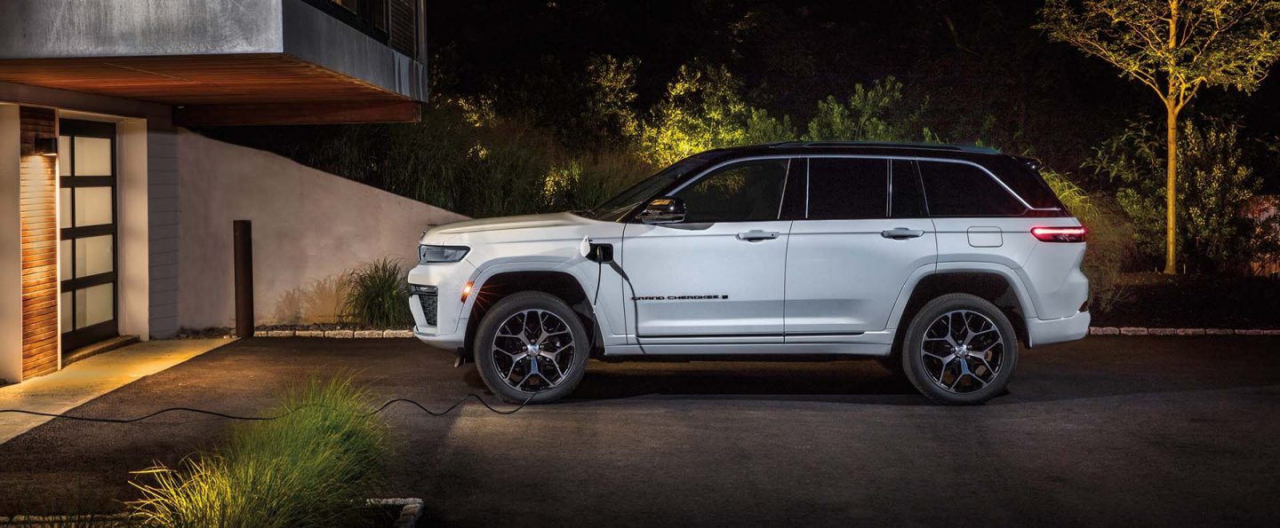 A driver-side profile of a white 2026 Jeep Grand Cherokee Summit 4xe parked in the driveway of a large home with a charging cord plugged into the vehicle's charging port.