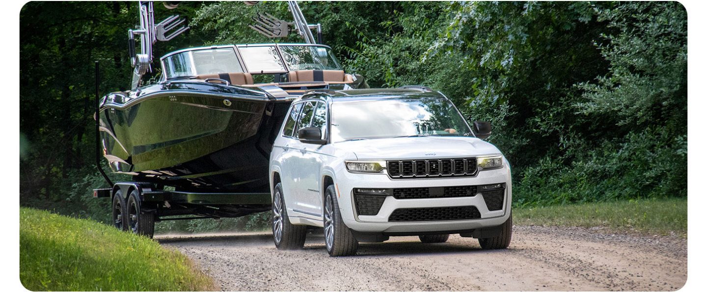 A white 2026 Jeep Grand Cherokee Summit traveling down a dirt road towing a large motorboat.
