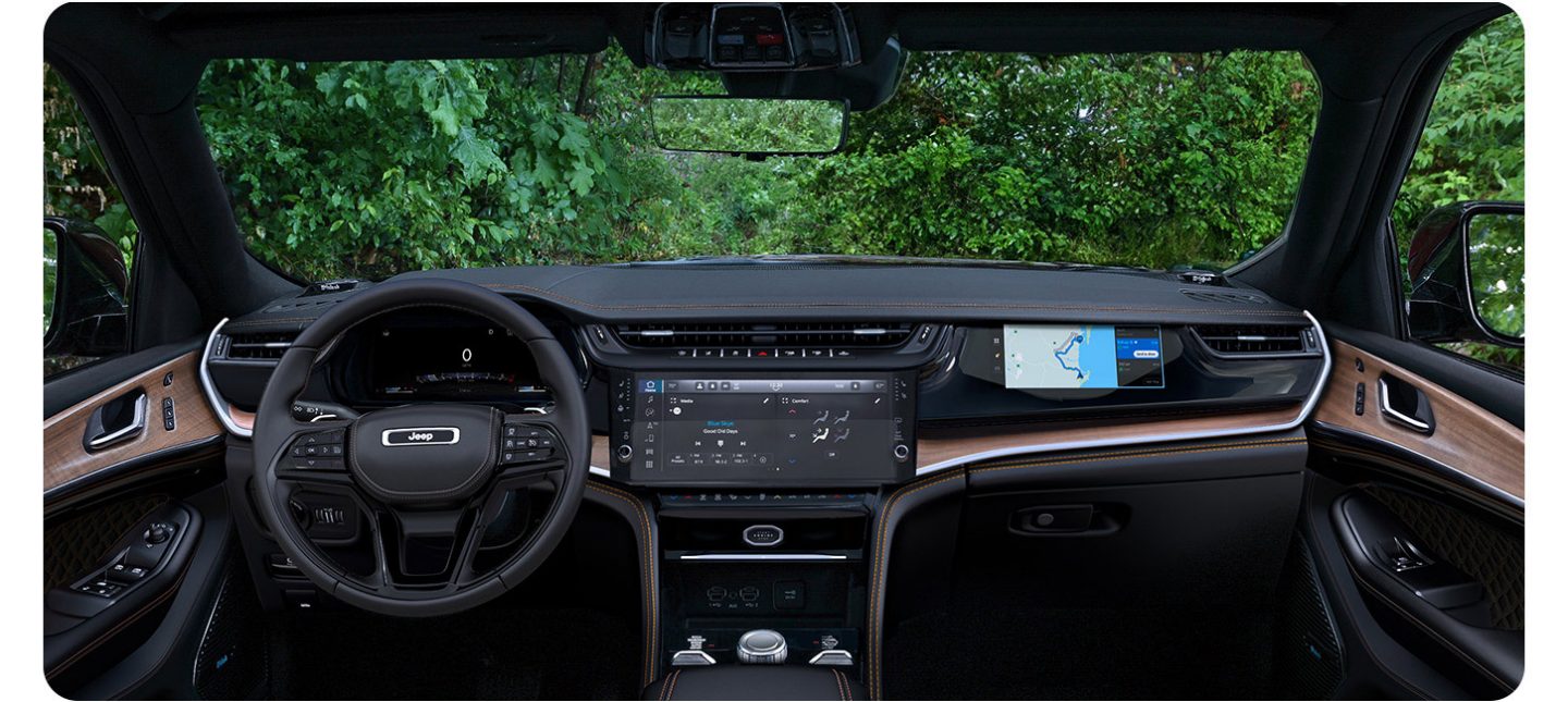 The front seat area of the 2026 Jeep Grand Cherokee Summit focusing on the steering wheel, Uconnect touchscreen and Front Passenger Interactive Display.