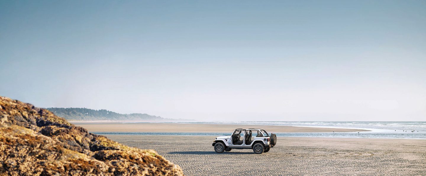 A white 2026 Jeep Wrangler Sahara four-door, with its doors and top removed, parked on a beach.