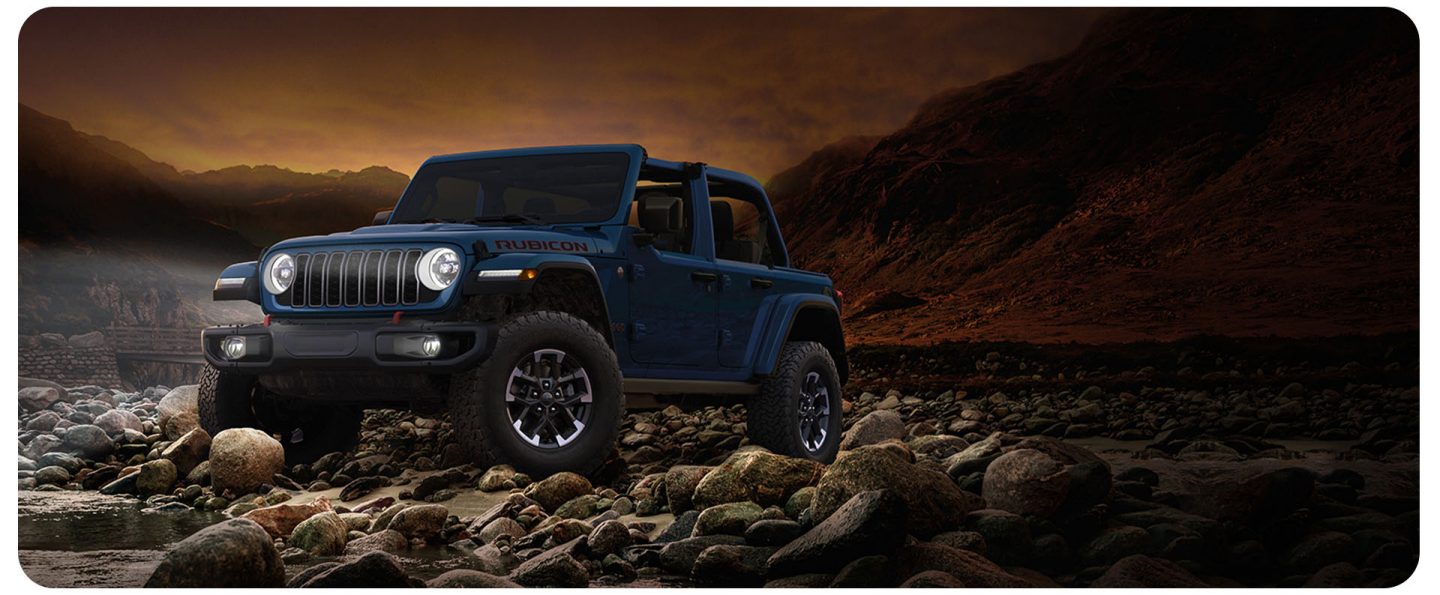 A blue 2026 Jeep Wrangler Rubicon X four-door, with its top removed and its headlamps on, parked on a rocky shoreline at the water's edge at night.