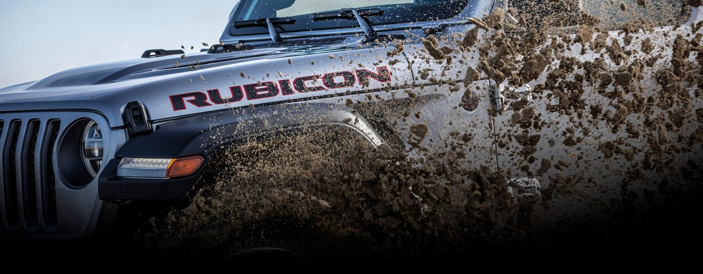 A close-up of a 2023 Jeep Gladiator Rubicon kicking up dirt and mud on its driver-side wheel and door.