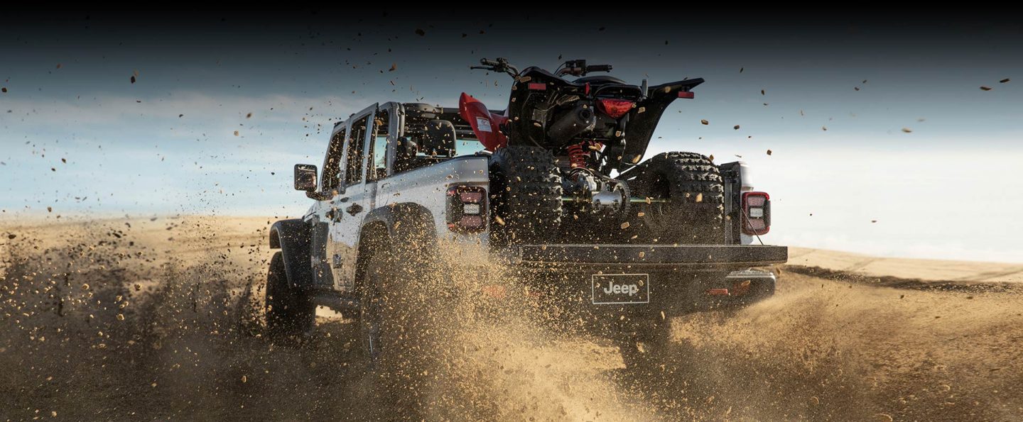 The rear view of a 2023 Jeep Gladiator Rubicon kicking up sand as it is driven across a desert, carrying an ATV in its bed with the tailgate open.