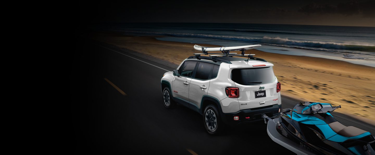 A white 2023 Jeep Renegade Trailhawk with a surfboard on its roof rack and a jet ski in tow.