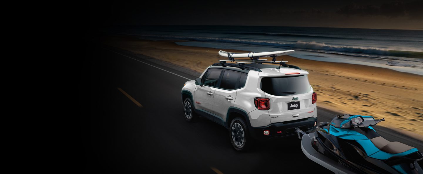 A white 2023 Jeep Renegade Trailhawk with a surfboard on its roof rack and a jet ski in tow.