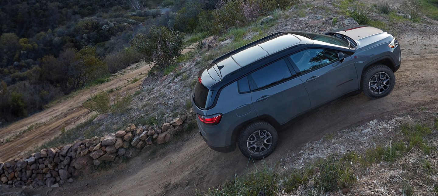 A 2024 Jeep Compass Trailhawk ascending a steep dirt road beside a stone fence.