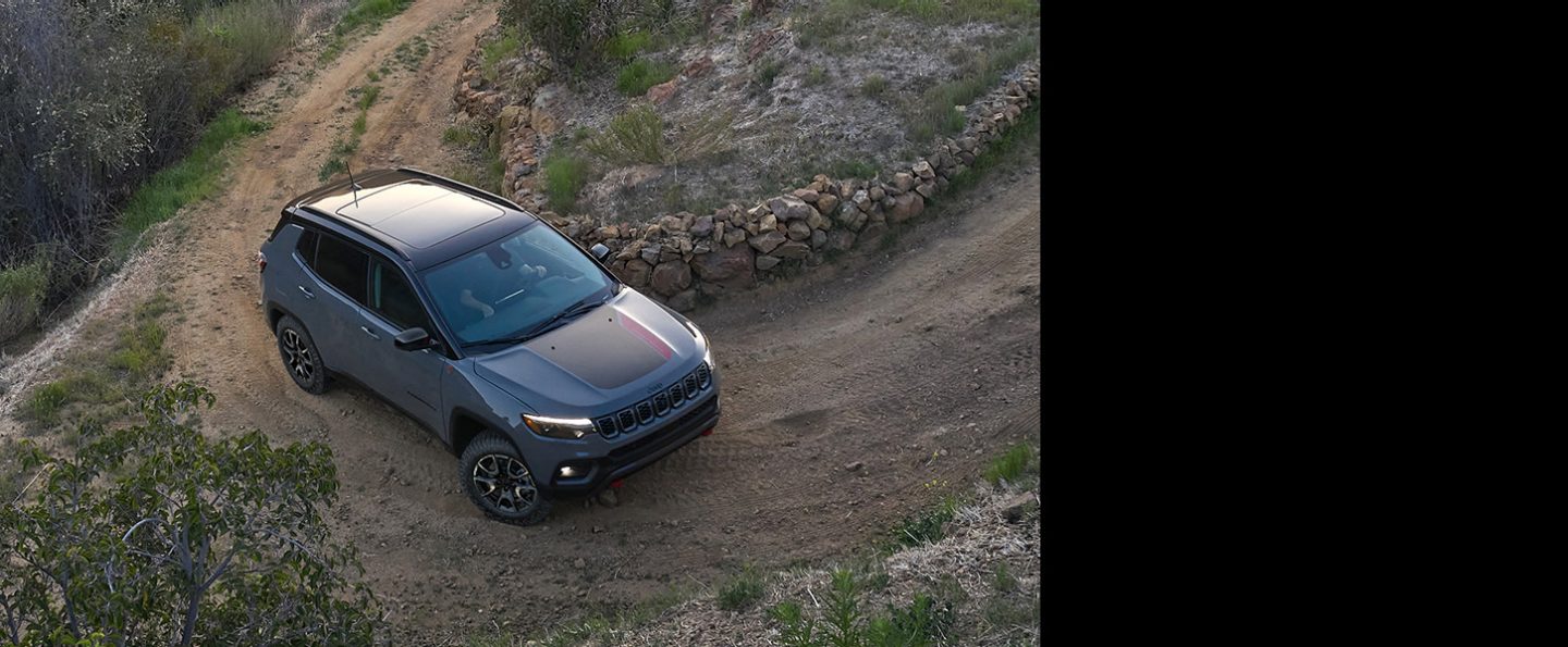 A 2024 Jeep Compass Trailhawk being driven on the tight curve of a dirt road hill beside a stone fence.