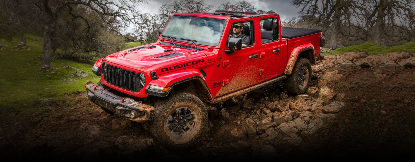 A red 2024 Jeep Gladiator Rubicon with its roof removed, being driven over a rocky, muddy trail off-road.