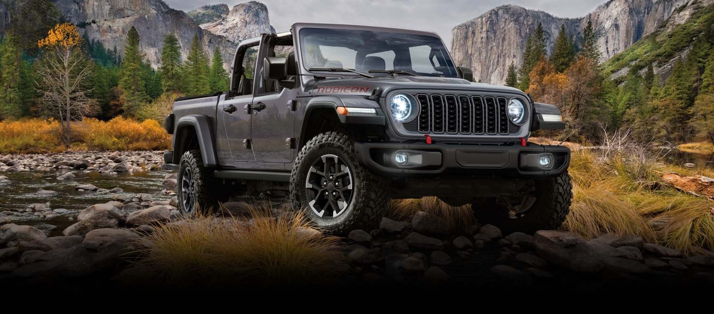 A gray 2024 Jeep Gladiator Rubicon exiting a stream off-road, with mountains in the background.