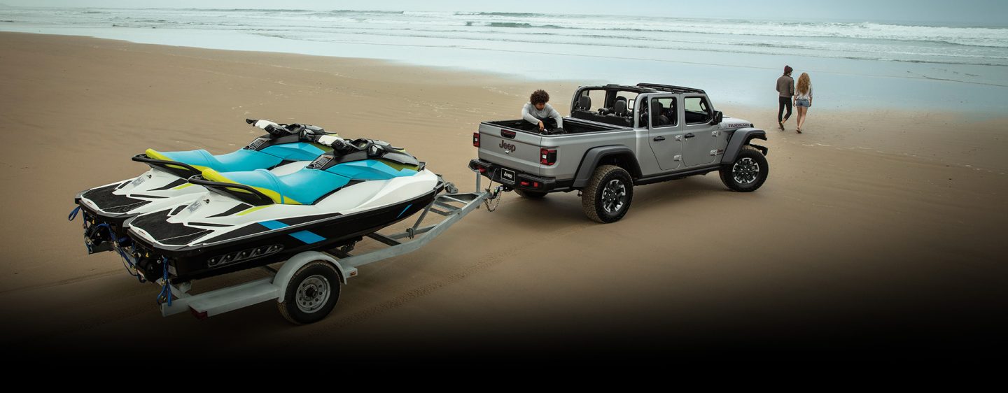 A silver 2024 Jeep Gladiator Rubicon with its roof removed, parked on a sandy beach near the water's edge, towing two personal watercrafts.