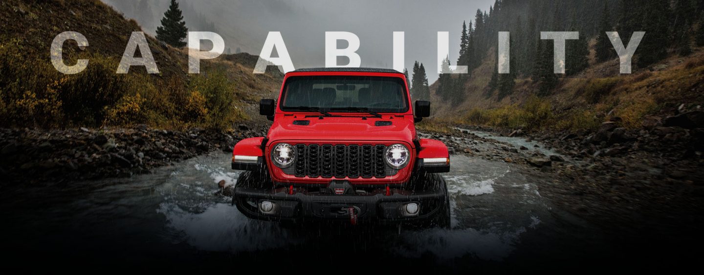 A front angle of a red 2024 Jeep Wrangler Rubicon being driven through a stream, following its path. Capability.
