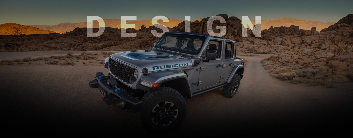 An angled driver-side profile of a gray 2024 Jeep Wrangler Rubicon 4xe parked on a clearing in the desert. Design.