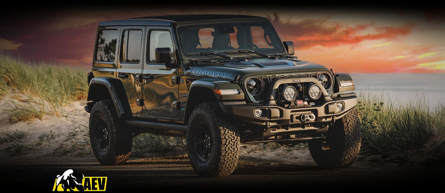 A 2024 Jeep Wrangler Rubicon 4xe with the Level II Package by American Expedition Vehicles parked beside a marshy beach at sunset. The AEV logo.